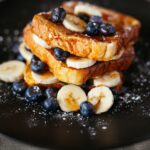 French toast topped with banana slices, blueberries, and syrup on a black plate.