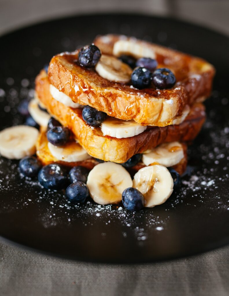 French toast topped with banana slices, blueberries, and syrup on a black plate.