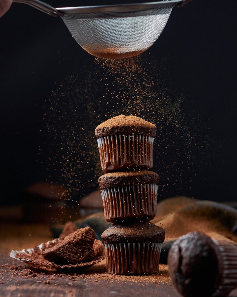 A stack of three chocolate muffins being dusted with cocoa powder.