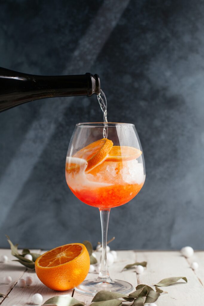 A cocktail glass filled with ice, orange slices, and red liquid being topped with a sparkling drink.