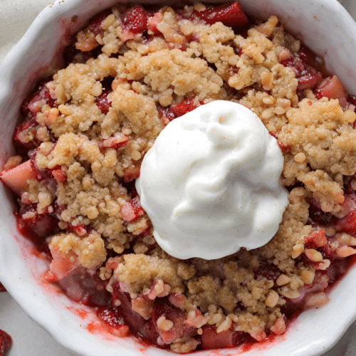 A close-up image of a freshly baked strawberry rhubarb crisp, showing the golden-brown crisp topping layered over the bubbling fruit filling in a white ceramic baking dish.