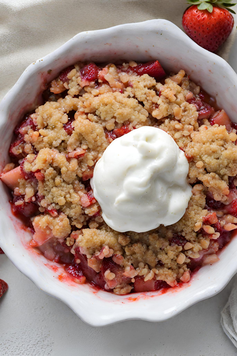 A close-up image of a freshly baked strawberry rhubarb crisp, showing the golden-brown crisp topping layered over the bubbling fruit filling in a white ceramic baking dish.