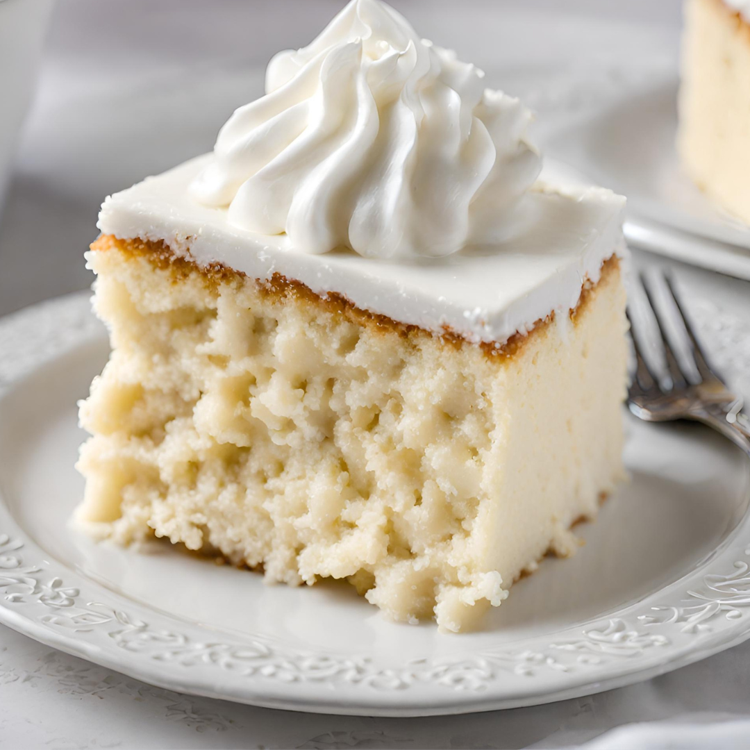 A slice of Tres Leches Cake on a white ornate plate, topped with a generous swirl of whipped cream. The cake has a moist texture visible from the soaked layers, showcasing a rich and creamy consistency.