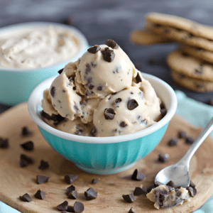 Scoops of cookie dough ice cream in a turquoise bowl garnished with chocolate chips, on a wooden board next to a spoon and cookies.