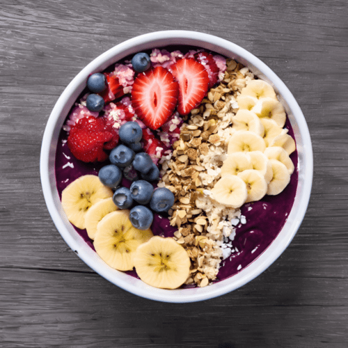 A colorful acai bowl in a white bowl, topped with fresh strawberries, blueberries, raspberries, banana slices, and granola, set on a gray wooden surface.