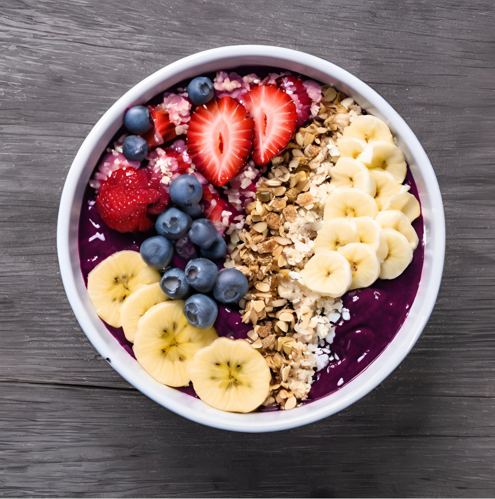 A colorful acai bowl in a white bowl, topped with fresh strawberries, blueberries, raspberries, banana slices, and granola, set on a gray wooden surface.