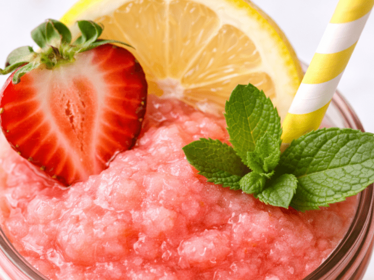Macro close-up of a strawberry lemonade slushie topped with a lemon slice, strawberry half, mint, and a yellow-striped straw.