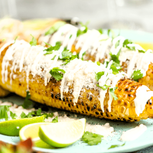 Grilled Mexican Street Corn (Elotes) topped with creamy sauce, crumbled cotija cheese, and fresh cilantro, served with lime wedges on a green plate.