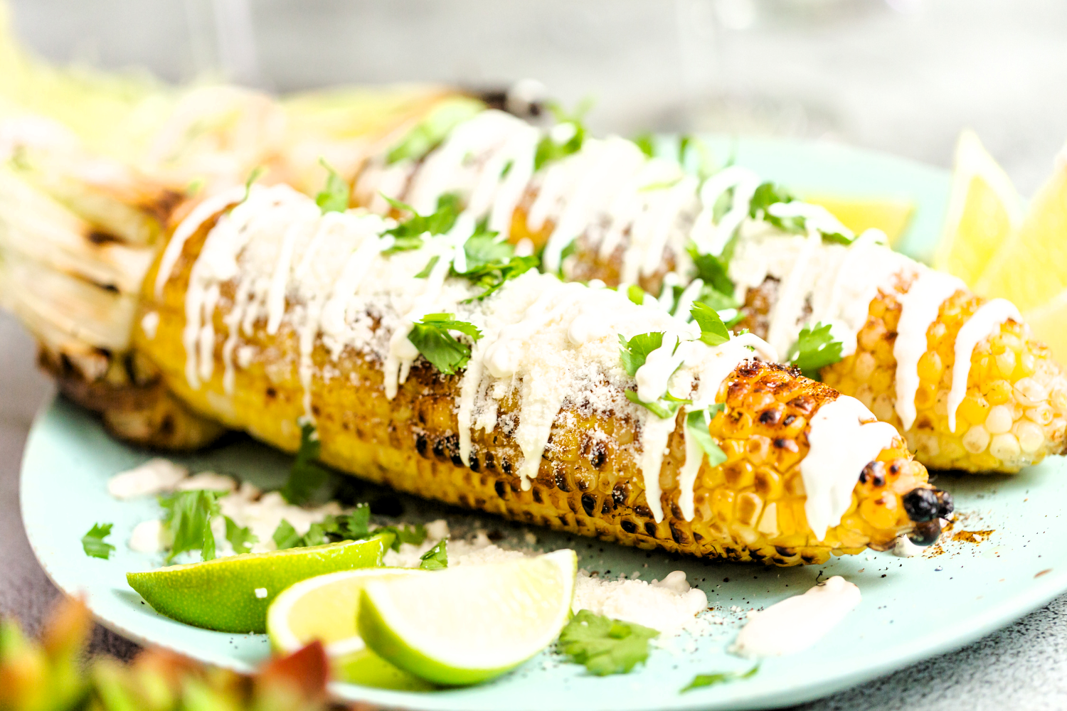 Grilled Mexican Street Corn (Elotes) topped with creamy sauce, crumbled cotija cheese, and fresh cilantro, served with lime wedges on a green plate.