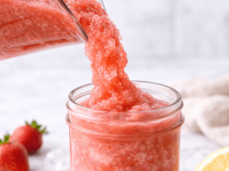 Strawberry lemonade slushie being poured into a mason jar on a marble counter with strawberries, lemon half, and ice.