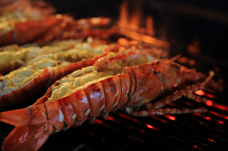 Close-up of succulent lobster tails grilling over an open flame, basted with garlic herb butter for a rich and flavorful summer BBQ dish. Find the full recipe on Satisfy Your Cravings. #GrilledLobster #SeafoodRecipes #SummerBBQ #SatisfyYourCravings