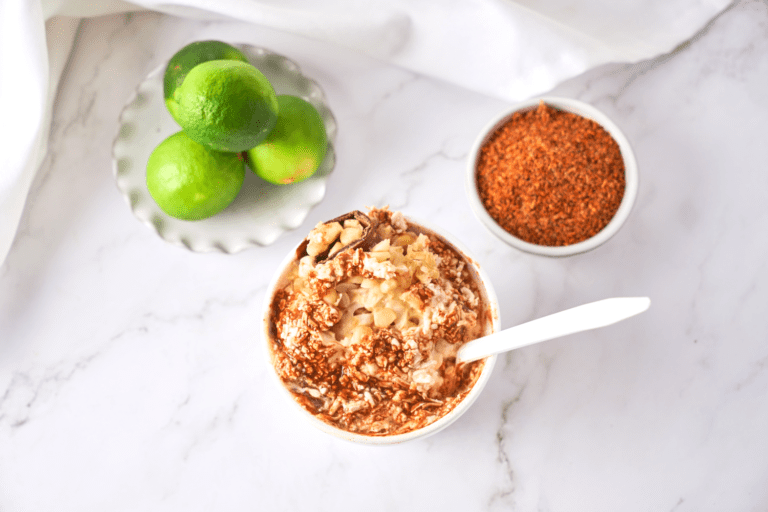 Mexican Street Corn in a cup topped with crumbled cheese and chili powder, accompanied by fresh limes and a bowl of seasoning on a marble surface. Perfect for enjoying the flavors of Elotes in a convenient and portable form.