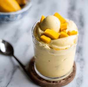 Gourmet mango frozen yogurt in a glass, topped with a scoop and diced mango pieces on a marble countertop.