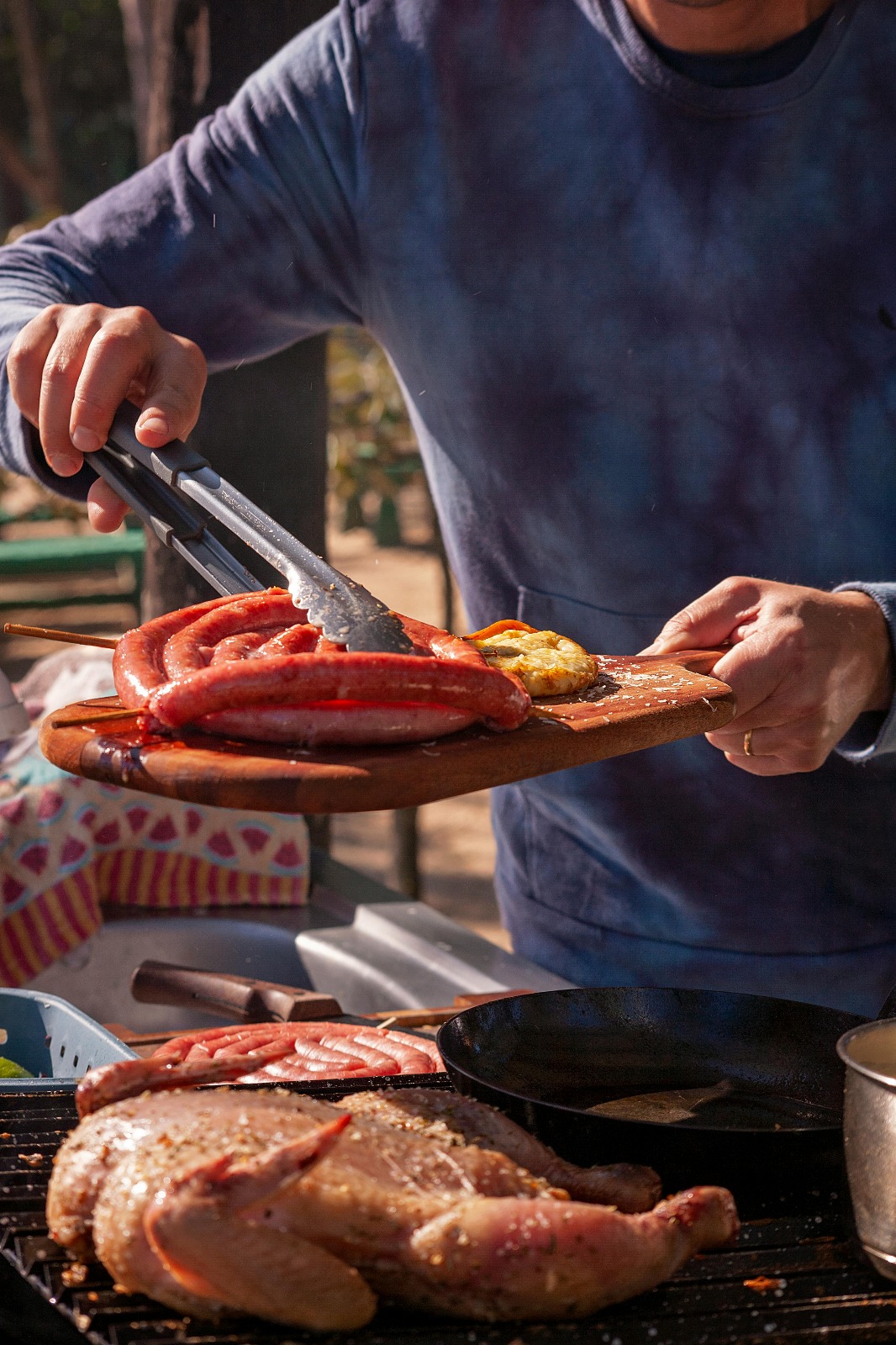 A person in a blue sweatshirt uses tongs to serve grilled sausages and vegetables onto a wooden platter at an outdoor barbecue.