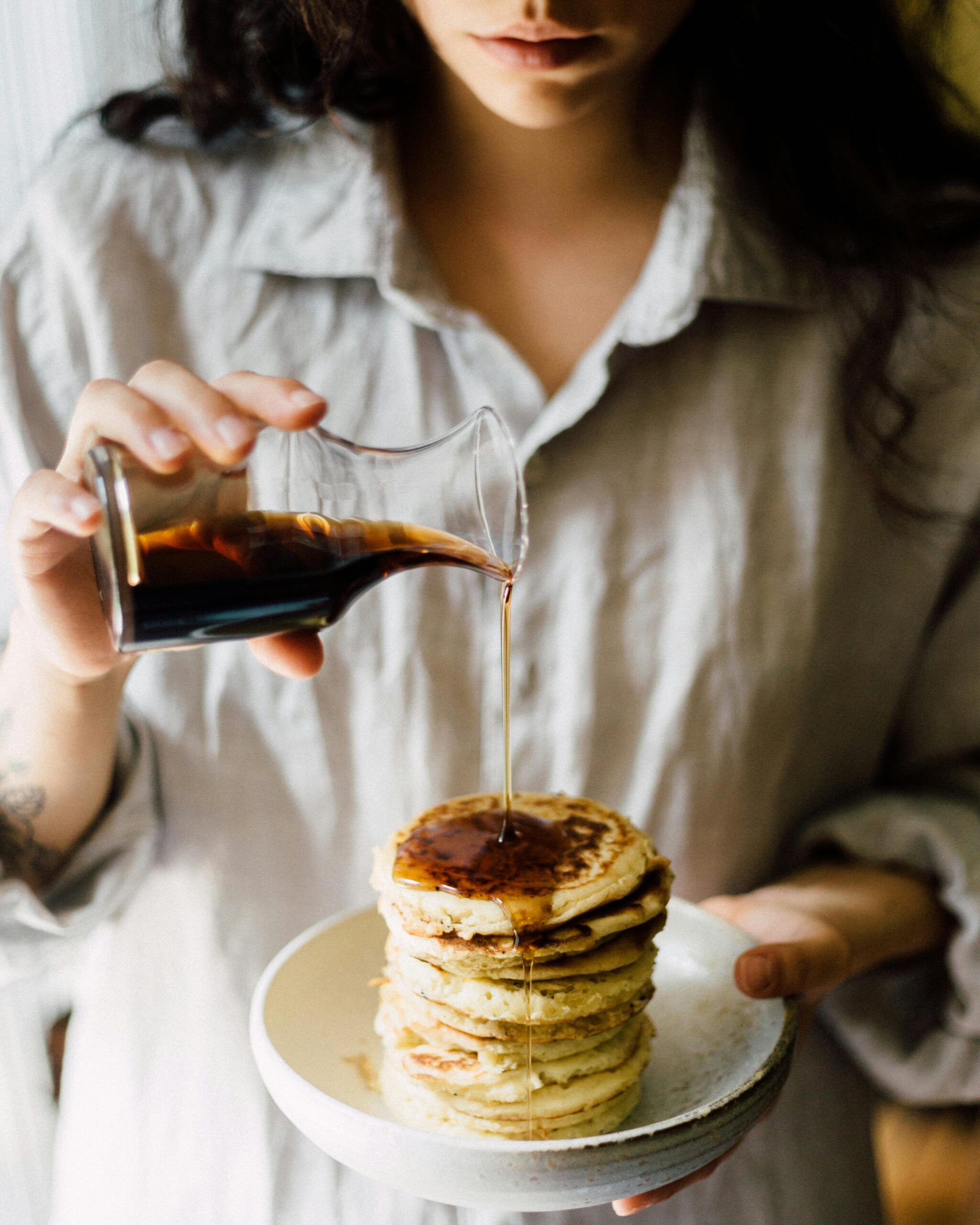 A woman in a linen shirt pours maple syrup over a tall stack of fluffy pancakes on a ceramic plate.