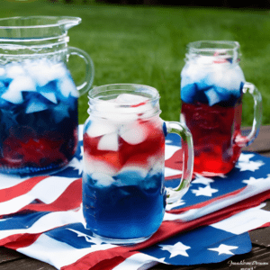 Festive Patriotic Punch for 4th of July - Red, White, and Blue Layered Drink in Clear Pitchers and Glasses with Ice and Straws on a Star-Spangled Tablecloth - Perfect for Summer Celebrations - Satisfy Your Cravings Blog