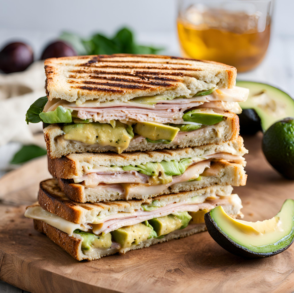 Stack of Turkey and Avocado Panini slices with cheese and fresh avocado on a cutting board.