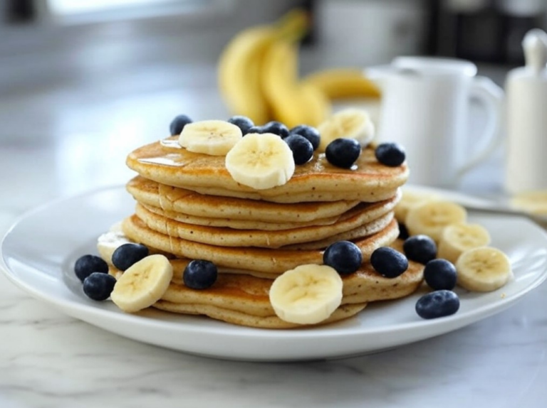 A close-up of fluffy banana oatmeal pancakes topped with bananas, blueberries, and maple syrup on a white plate.