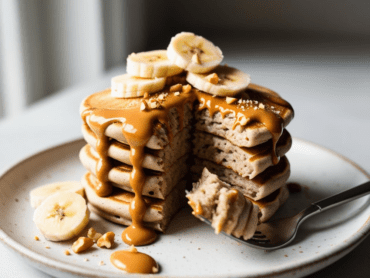 A cut stack of peanut butter banana protein pancakes with a peanut butter drizzle, banana slices, and a fork with a bite-sized piece.