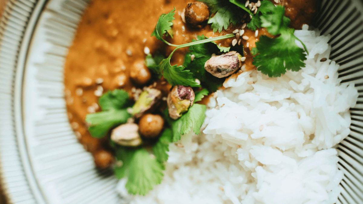 Close-up of creamy curry with chickpeas served alongside white rice, topped with cilantro, pistachios, and sesame seeds.